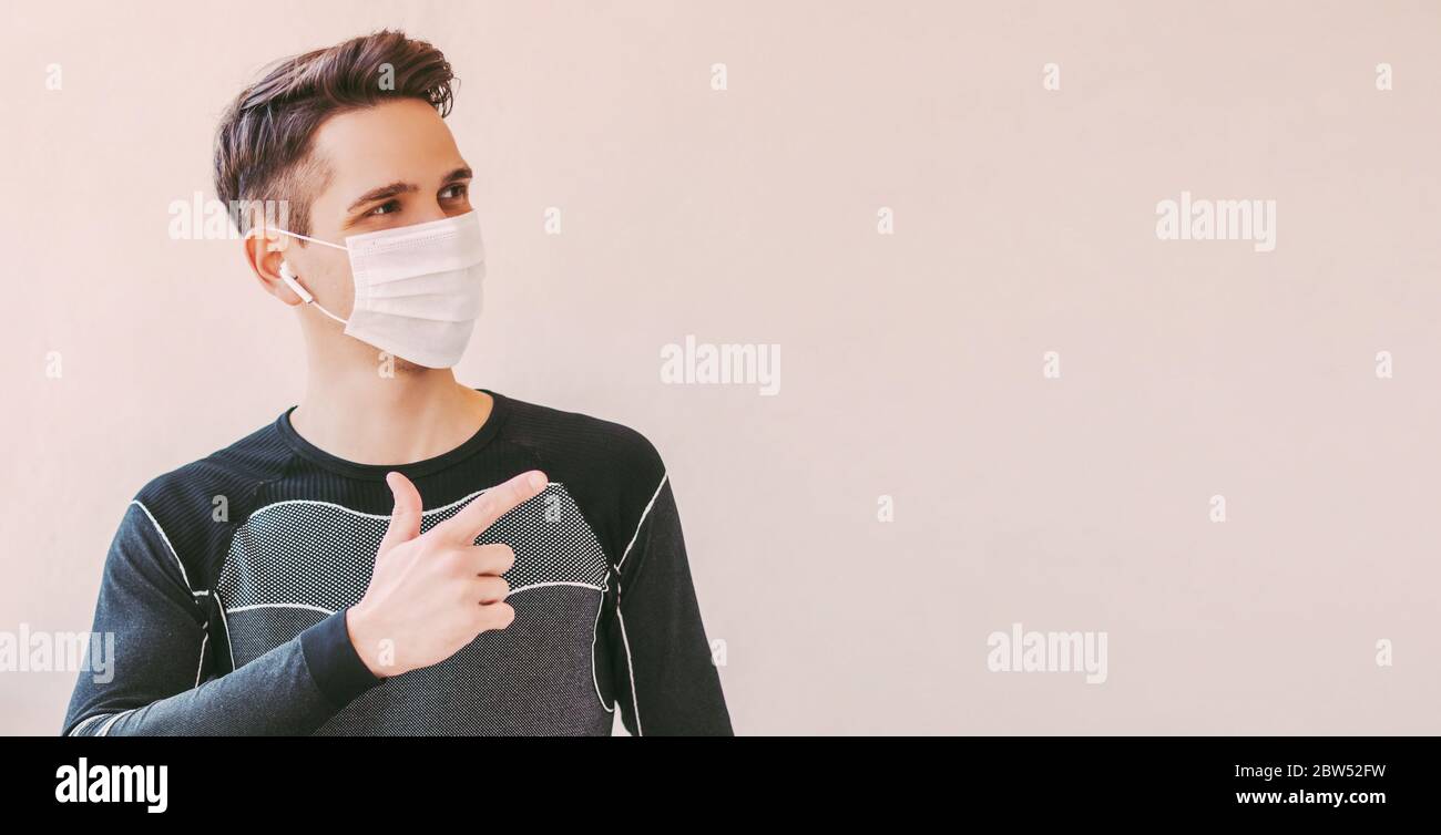 Junge zuversichtlich Sportmann in medizinischen Gesichtsmaske zeigt mit Finger auf Hintergrund mit Kopierraum. Happy Fitness Trainer in Schutzmaske zeigt c Stockfoto