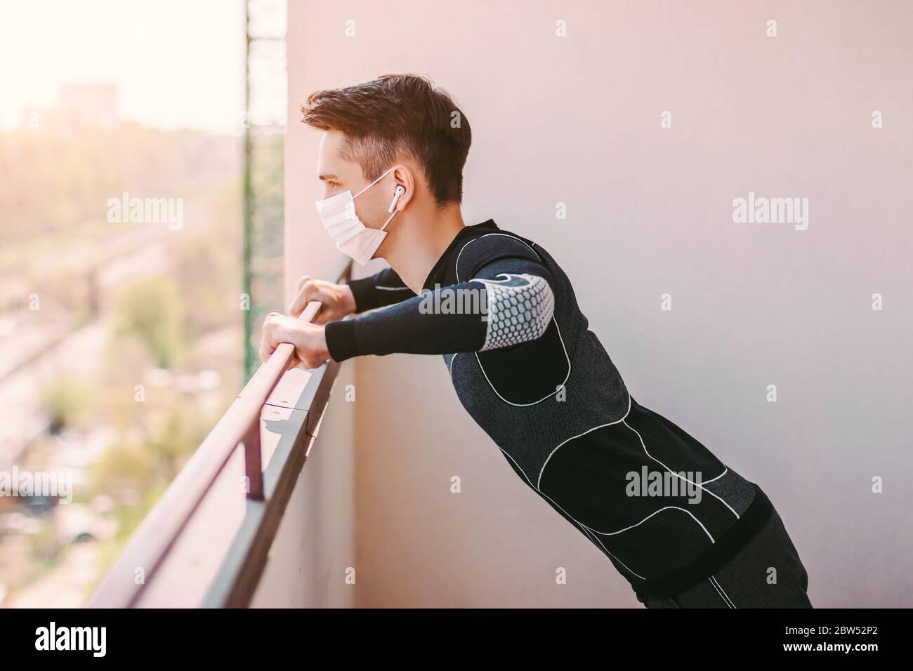 Sportlicher junger Mann Fitness-Trainer in medizinischen Gesichtsmaske tun Liegestützübungen auf dem Balkon. Selbstbewusste Sportmann in Schutzmaske Training worko Stockfoto