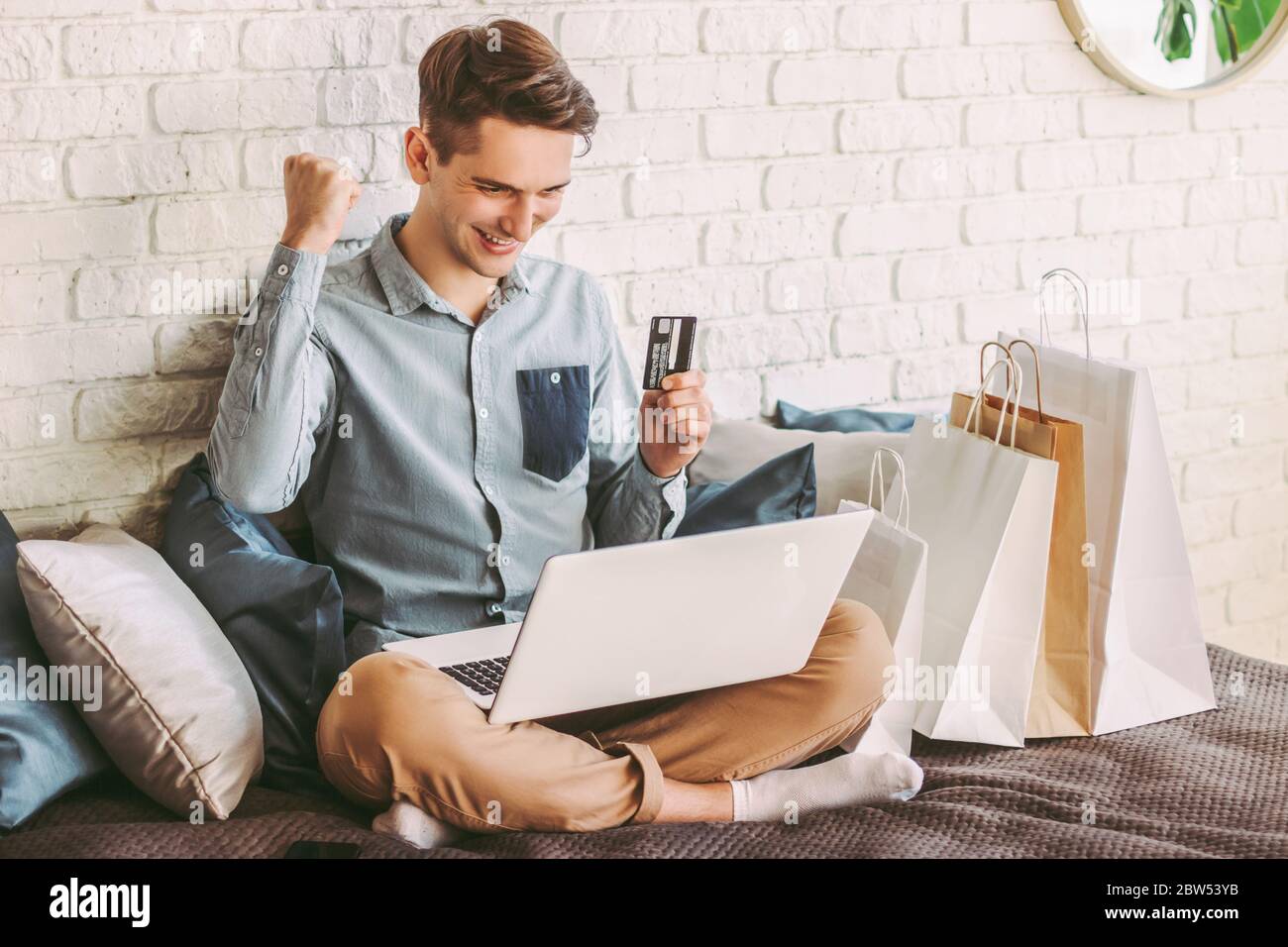 Glücklich männliche Shopper online einkaufen mit Laptop, halten Sie Kreditkarte in der Hand und lächelnd auf der Couch. Junger stilvoller Hipster-Mann feiert erfolgreichen Kauf Stockfoto