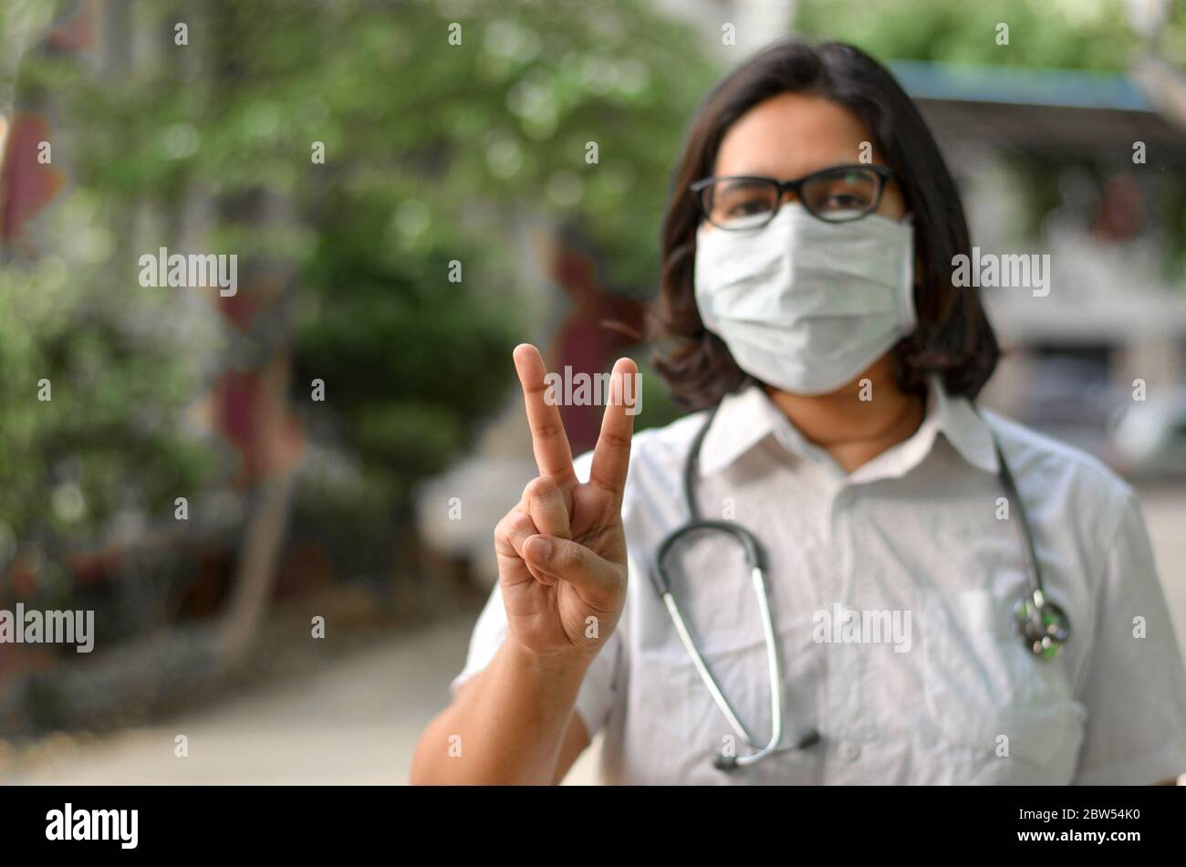Porträt einer jungen medizinischen Arbeiterin, die mit ihrer rechten Hand ein Siegeszeichen macht und eine chirurgische Maske trägt, um sich vor Corona VI zu schützen Stockfoto