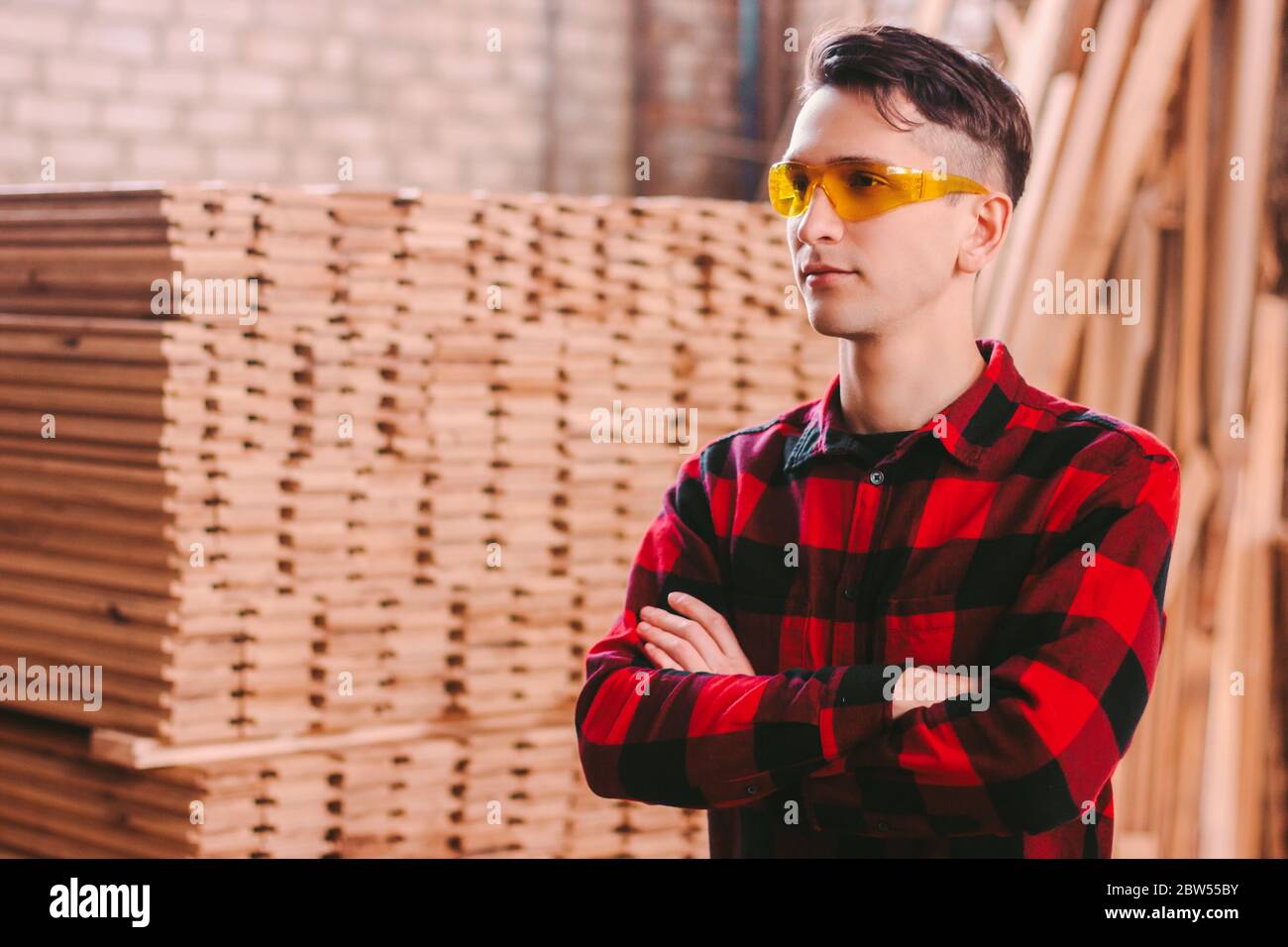 Selbstbewusste männliche Schrankbauer in Schutzbrille halten die Arme gekreuzt, während am Sägewerk stehen. Junger Zimmermann in der Holzwerkstatt. Kleines Holzprodu Stockfoto