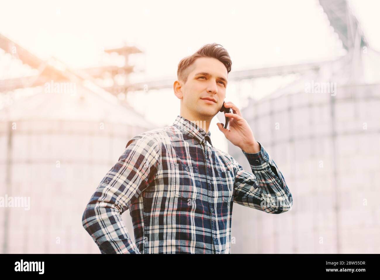 Porträt des jungen Geschäftsmanns, der während der Arbeit in der Silagelagerfabrik Handy spricht. Glücklich Landwirtschaft Business Manager hält Handy in Stockfoto