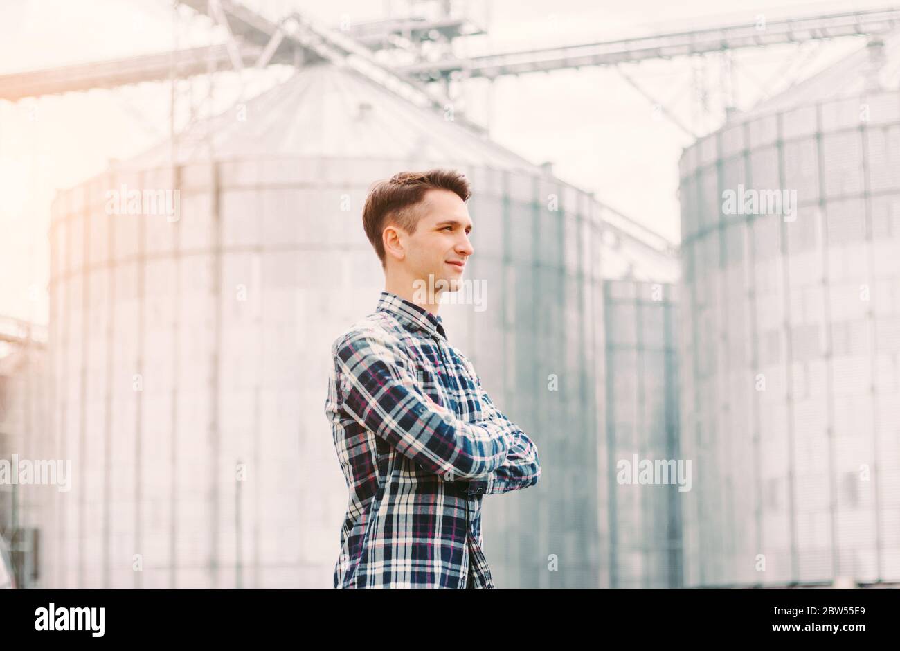 Porträt des selbstbewussten Geschäftsmann Besitzer von Getreideanlagen halten die Arme gekreuzt, während gegen Silos Gebäude stehen. Junge erfolgreiche Agricultu Stockfoto