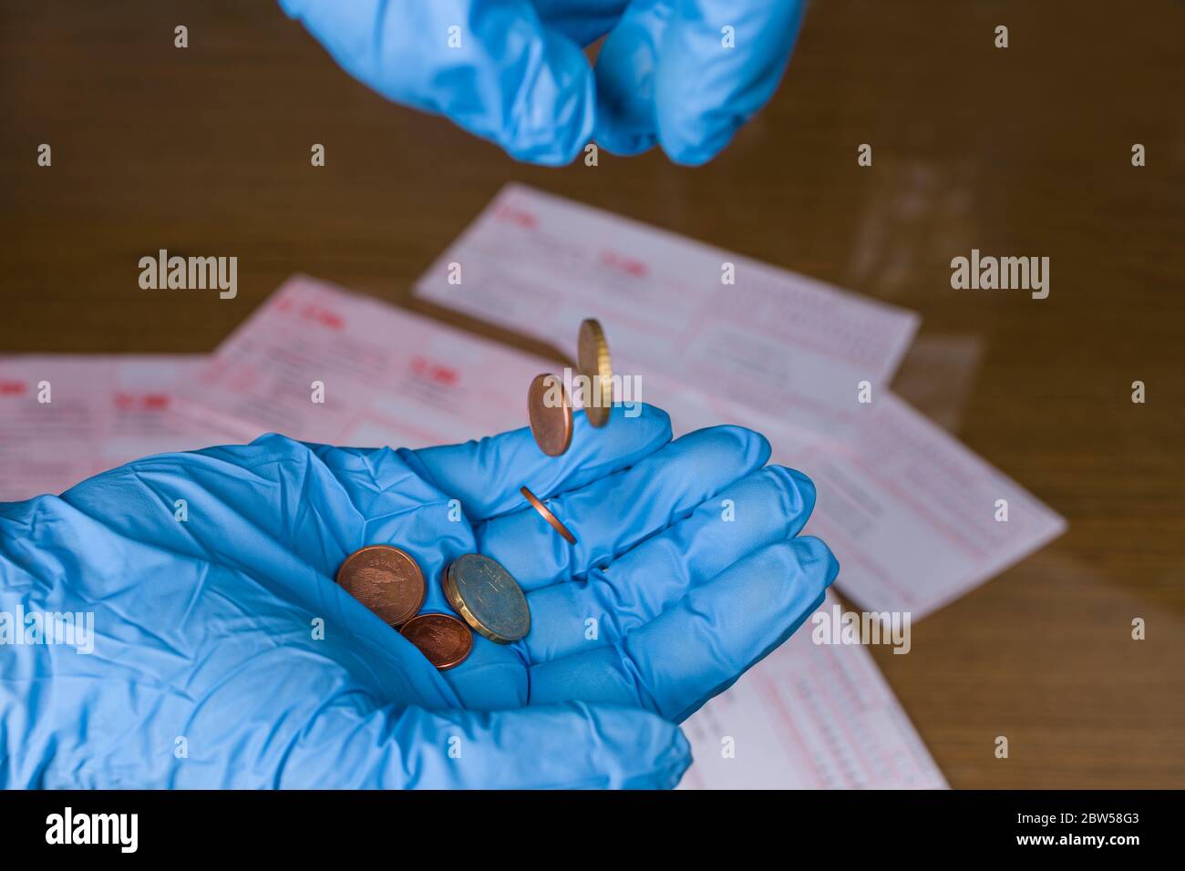 Euro-Münzen fallen in blauen Schutzhandschuh auf die Handfläche, Geldbestellungen auf Holzschreibtisch. Wirtschaftlicher und finanzieller Verlust. Armut Not in Coronavirus Pandemie. Stockfoto