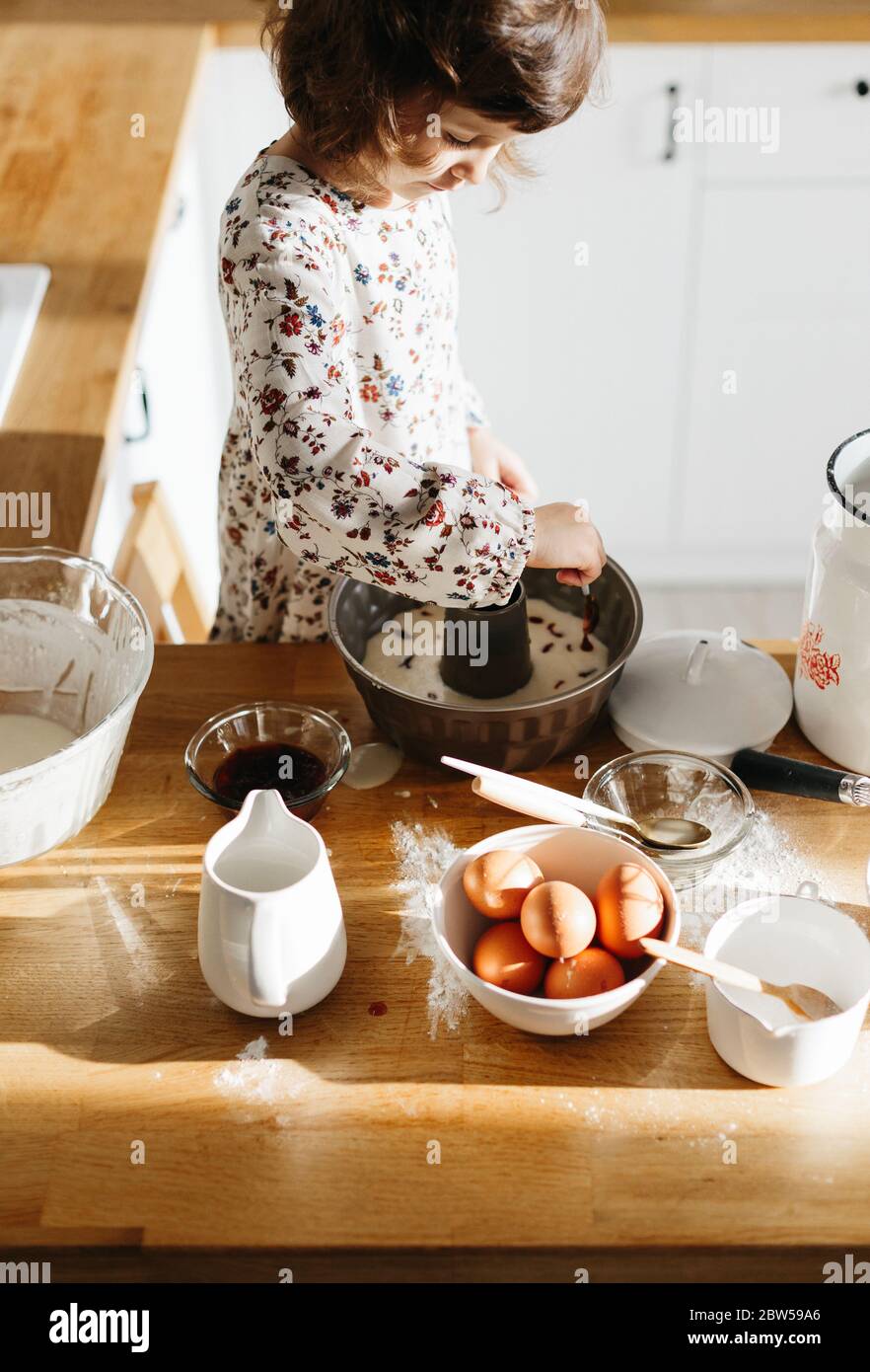 Cute 5 Jahre Mädchen tragen Kleid Vorbereitung Kuchen in der weißen modernen Küche. Mit rohem Teig Konfitüre in die Backform geben Stockfoto