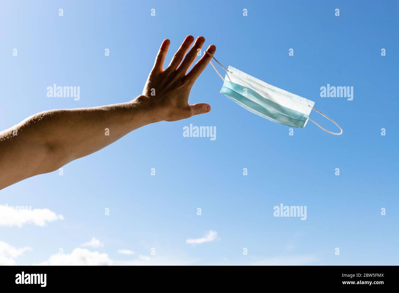 Ende der Quarantäne, Covid-19. Mann, der abzieht und wegwirft gebrauchte medizinische Schutzmaske hält, es auf ihrem Finger auf blauem Himmel Hintergrund, genießt das Leben, c Stockfoto