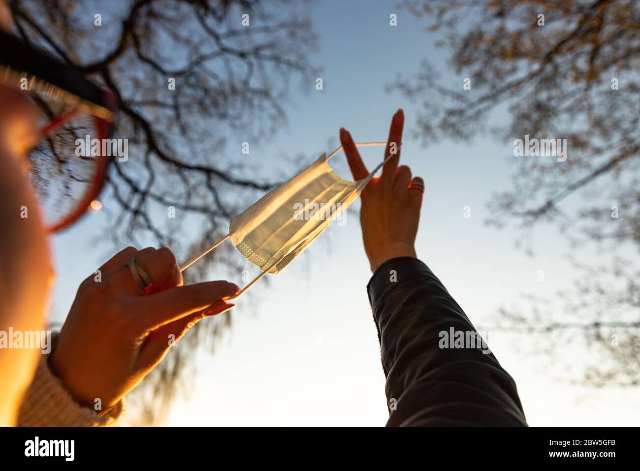 Frau verwendet eine schützende medizinische Maske als Schleuder bei Sonnenuntergang, selektiven Fokus auf Maske, im Freien. Genießt das Leben, zurück zum normalen Leben nach covid-19 pand Stockfoto