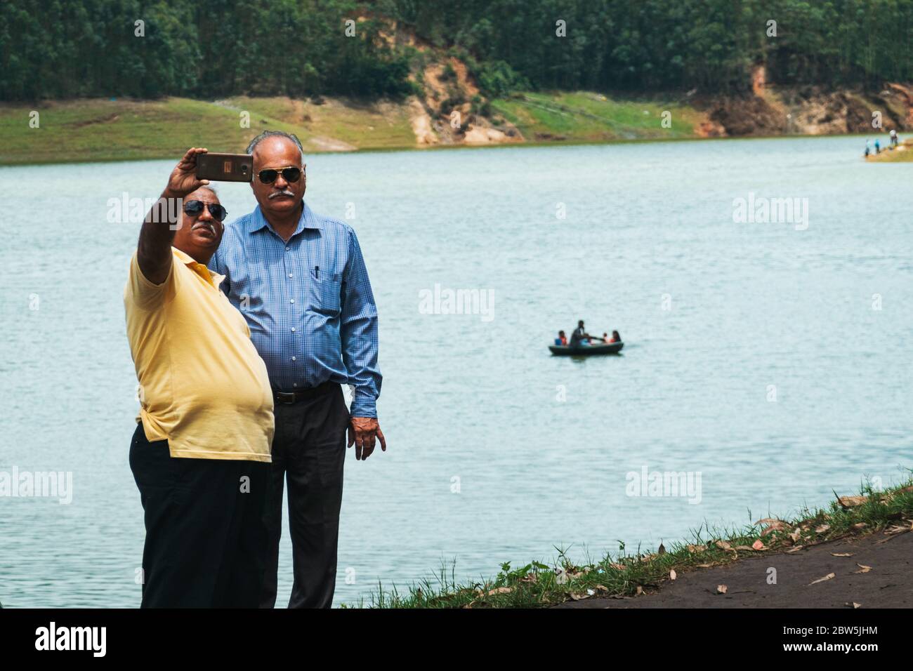 Zwei ältere Männer machen ein Selfie am Echo Point, auf dem Mattupatti-Staudamm in Munnar, Kerala, Indien. Hinter dem Hotel ruht ein Boot von Touristen Stockfoto