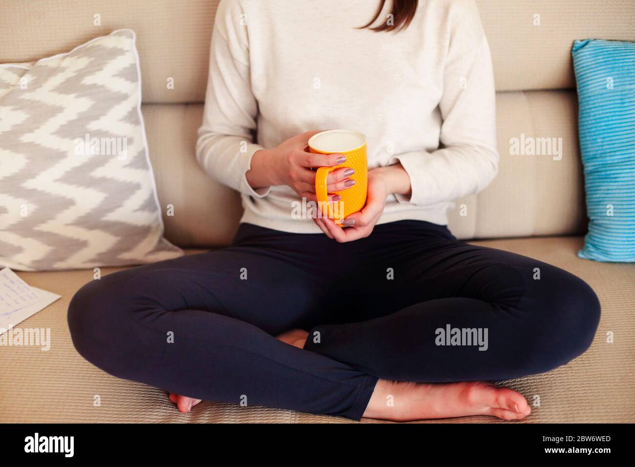 Schneiden Sie anonyme Frau in lässiger Kleidung sitzen auf einer bequemen Couch und genießen heißen Cappuccino während der Abkühlung zu Hause am Wochenende Stockfoto
