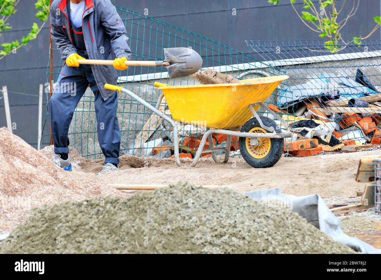 Ein Arbeiter in einem dunkelblauen Overall mit einer Schaufel lädt Sand in einer gelben Baukarre vor dem Hintergrund einer Baustelle. Stockfoto