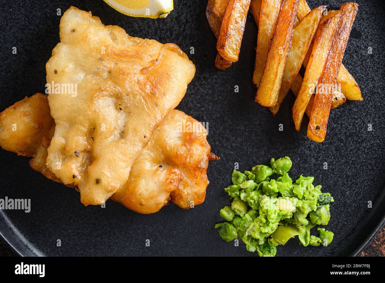 Englisch Traditional Fish and Chips with pürhed Minty Erbsen, Tartar Sauce vertikale Seitenansicht auf schwarzem Teller close up Details Seitenansicht. Überalt rustikal Stockfoto