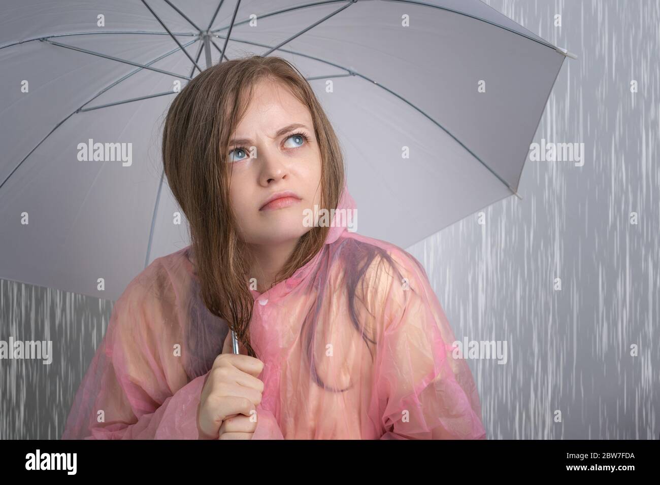 Junge kaukasische Frau Mädchen in einem rosa Regenmantel hält Regenschirm unter starkem Regen mit lustig verärgert Gesicht. Konzept der Regenzeit Stockfoto