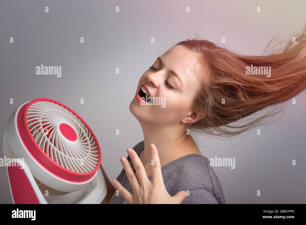 Junge kaukasische Frau Mädchen mit langen Haaren hält elektrischen Ventilator vor ihrem Gesicht. Kühlen Sie sich ab bei heißem Sommerwetter Stockfoto