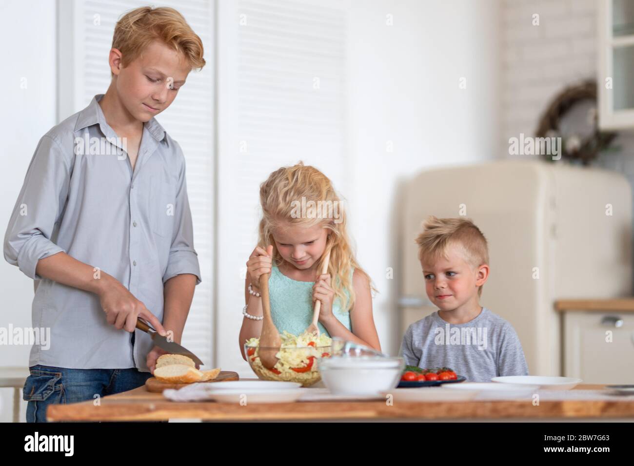 Drei blonde Kinder twoboys und Mädchen auf Küche zu Hause. Familie bereitet gesunde Lebensmittel, die Herstellung von grünem Salat. Stockfoto