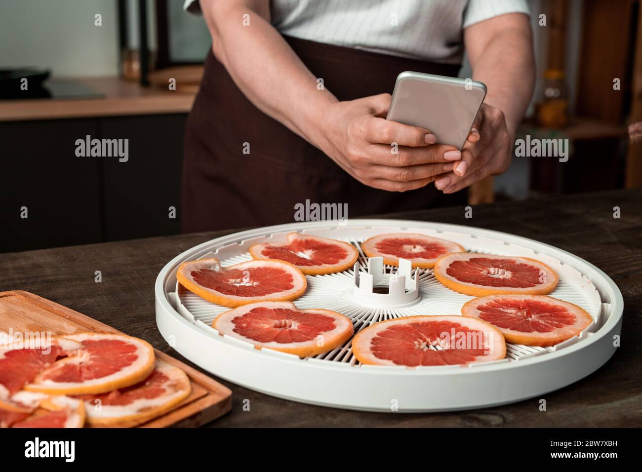 Hände der Hausfrau mit Smartphone über runden Tablett mit Obsttrockner mit frisch geschnittenen Grapefruit auf dem Tisch, während sie Foto von ihnen Stockfoto