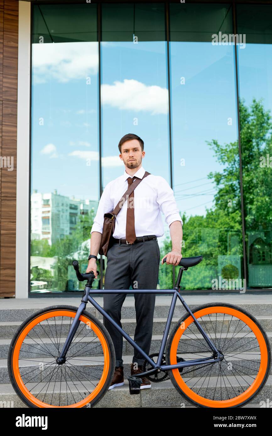 Junger eleganter Geschäftsmann in Hose, Hemd und Krawatte, der das Fahrrad hält, während er auf der Treppe gegen das Äußere des modernen Gebäudes steht Stockfoto