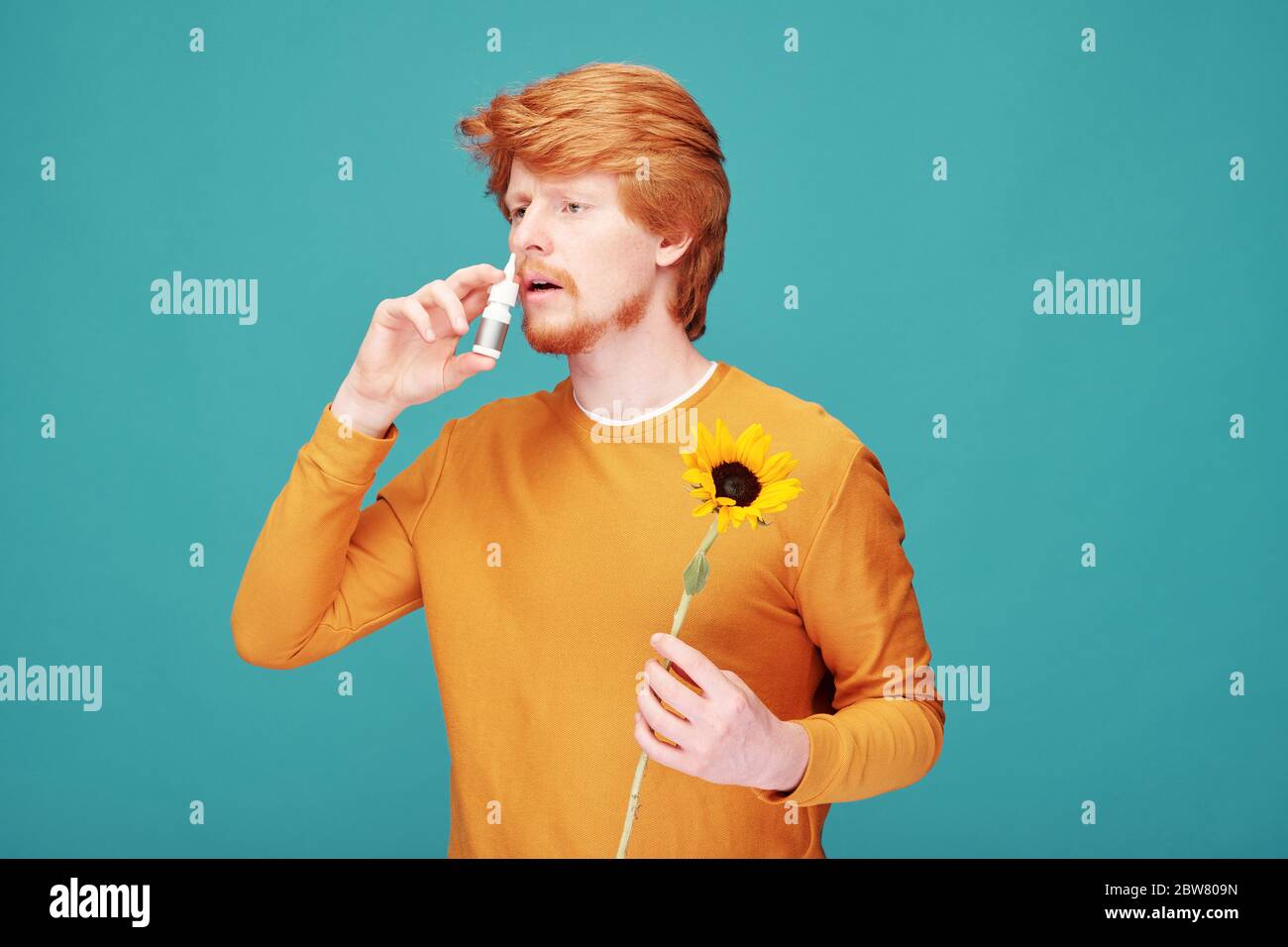 Junger Mann allergisch auf den Geruch von Sonnenblumen sprühen Anti-Allergie-Medizin in kleine Plastikflasche in seine Nase vor blauem Hintergrund Stockfoto