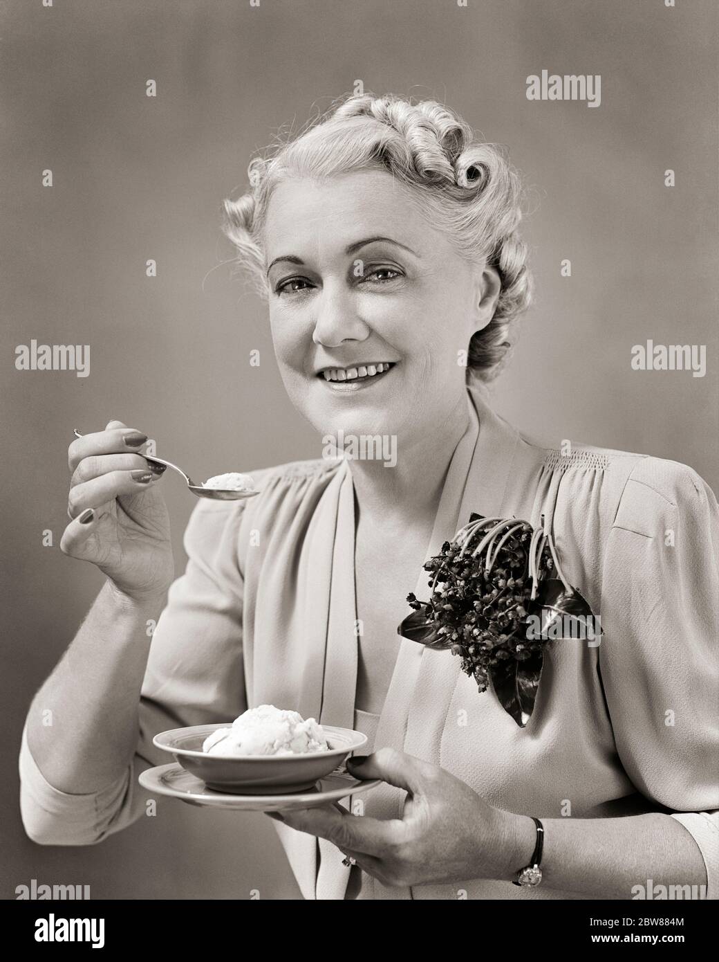 1940ER JAHRE FRAU MITTLEREN ALTERS ESSEN EIN GERICHT EIS TRAGEN KLEID MIT CORSAGE VON KÜNSTLICHEN BLUMEN LÄCHELND SUCHEN TAT KAMERA - F9727 HAR001 HARS LIFESTYLE ZUFRIEDENHEIT ÄLTERE FRAUEN STUDIO-SCHUSS GESUNDHEIT ZU HAUSE LEBEN KOPIE RAUM HALBE LÄNGE DAMEN PERSONEN VERTRAUEN MITTLEREN ALTERS B & W AUGENKONTAKT GLÜCK MITTLEREN ALTERS FRAU FRÖHLICH OLDSTER STILE ERNÄHRUNG LÄCHELN ÄLTESTEN KONSUMIEREN KONSUMIEREN CORSAGE FREUDIGE NAHRUNG STILVOLLE MODE KÜNSTLICHE SCHWARZ-WEISS KAUKASISCHEN ETHNIZITÄT HAR001 ALTMODISCHE TAT Stockfoto