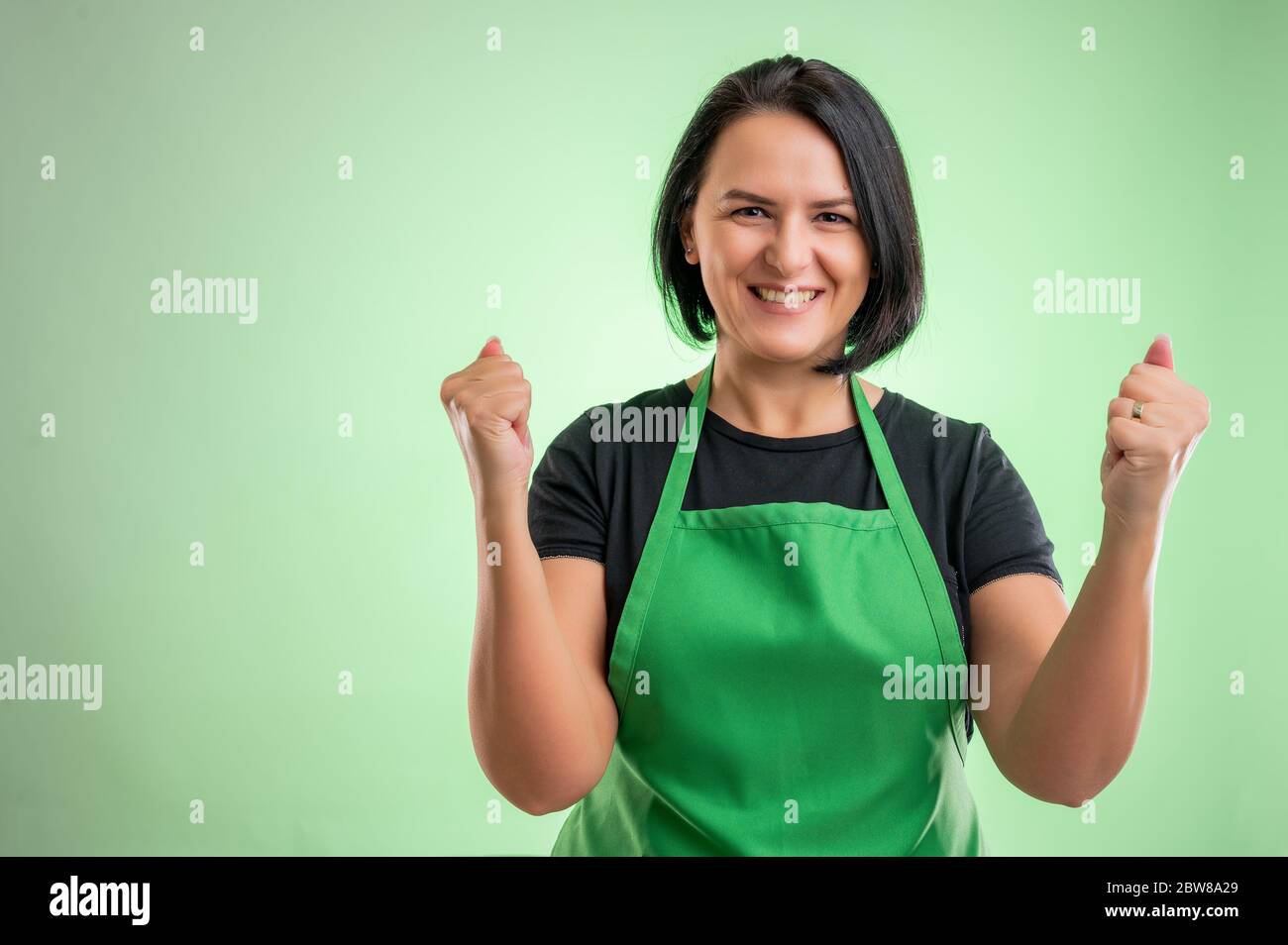 Köchin mit grüner Schürze und schwarzem T-Shirt, erhobene Fäuste, die mit Freude und Aufregung isoliert auf grünem Hintergrund ausrufen Stockfoto