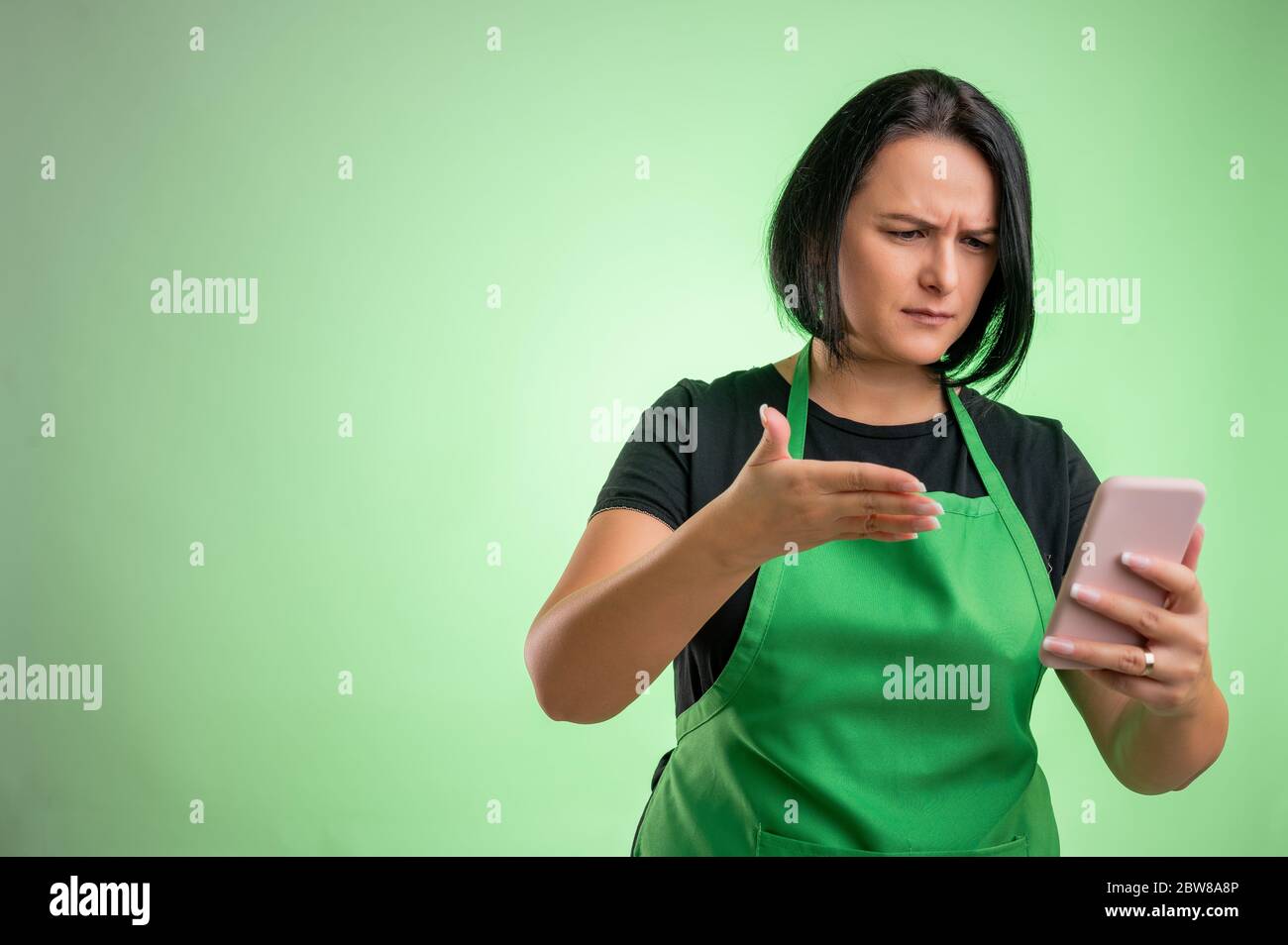 Köchin mit grüner Schürze und schwarzem T-Shirt, Smartphone haltend, SMS, nervös isoliert auf grünem Hintergrund Stockfoto