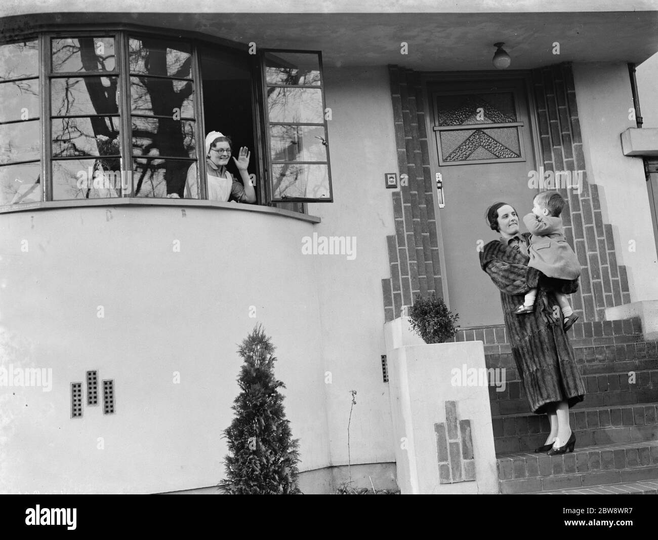 Frau Zehnder mit ihrem kleinen Jungen, an der Haustür ihres Hauses. Das Kindermädchen winkt ihnen vom Fenster auf Wiedersehen. Werbeaufnahmen für den Daily Mirror gemacht. 1937 Stockfoto