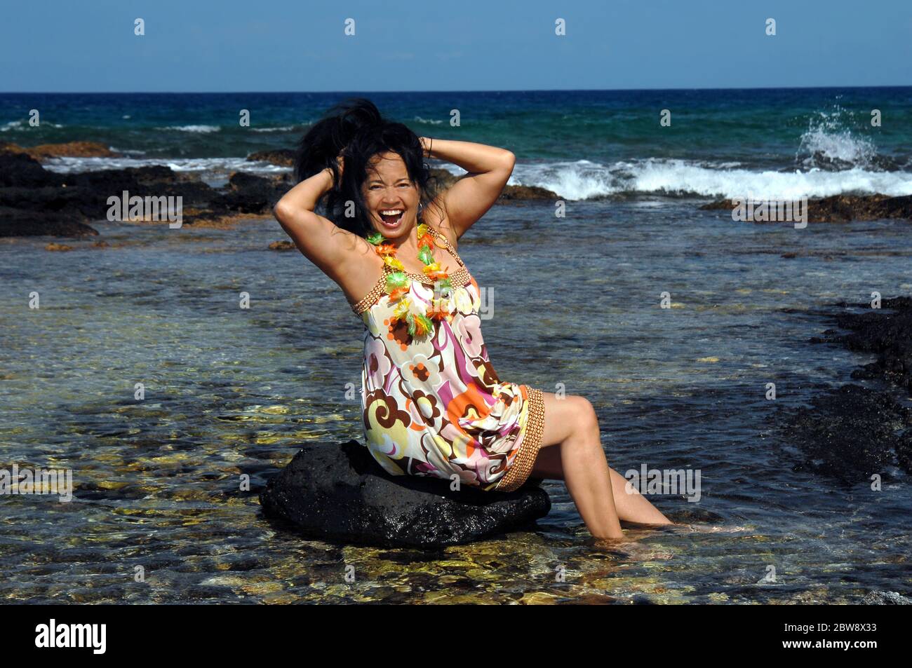 Hawaiianerin sitzt auf einem Felsen im flachen Wasser und feiert, indem sie ihre Haare hochwirft und posiert. Anaehomalu Bay an der Kohala Küste der Big Isla Stockfoto