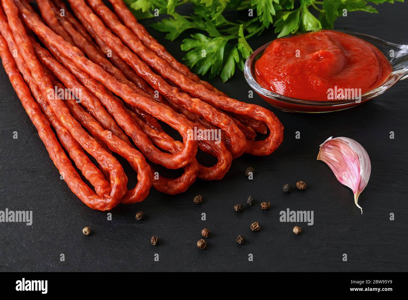Bund von dünnen trockenen geräucherten Wurst Kabanos oder Cabanossi und Tomaten Ketchup auf einem schwarzen Stein Servierbrett. Traditionelle polnische Fleischspezialitäten. Vorderansicht. Stockfoto