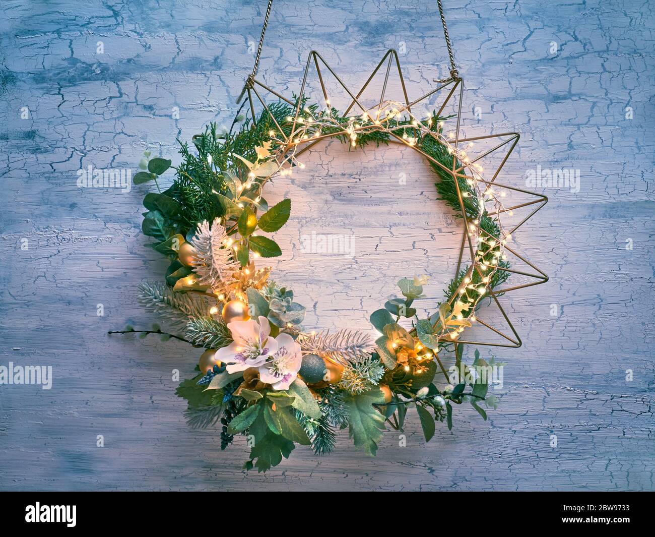 Dekorativer Weihnachtskranz mit Tanne, Winterpflanzen und Anemonblüten auf geometrisch vergoldetem Metallrahmen mit heller Girlande, die an einer Holztür hängt. Stockfoto