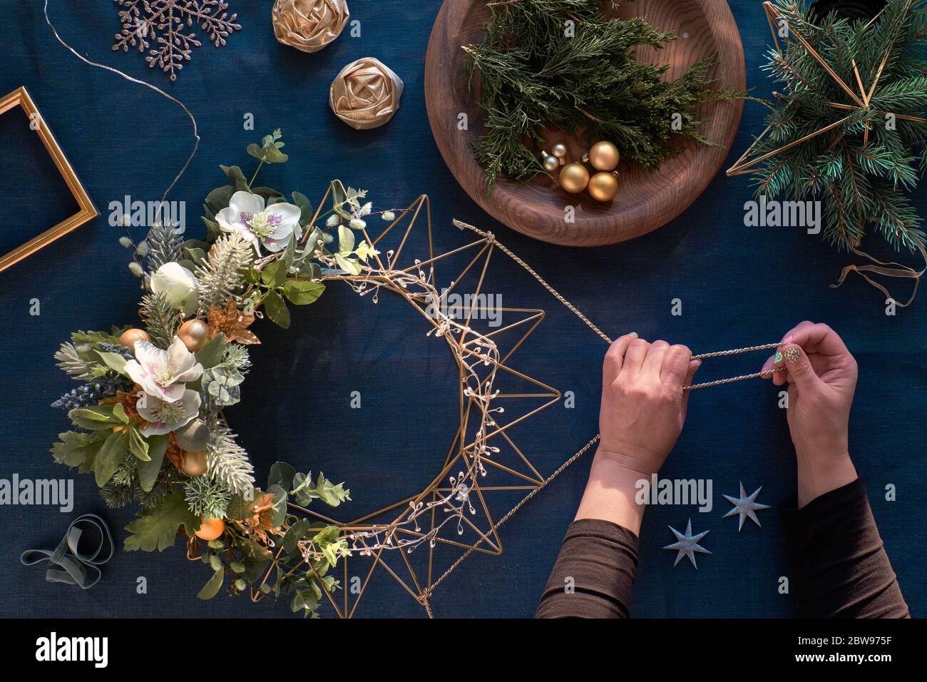 Herstellung von dekorativen Weihnachtskranz auf klassisch blauem Leinen. Frauenhände mit handgefertigtem Kranz. Weihnachtsschmuck, goldene Schmuckstücke, dekorative Anemone f Stockfoto