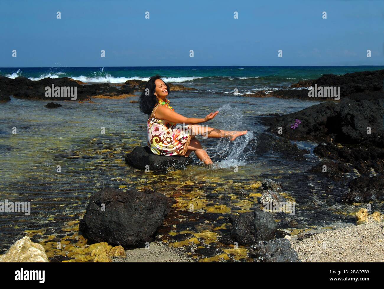 Ältere Hawaiiianerin wadet in die Untiefen und sitzt auf einem schwarzen Lavastein, der klatscht und spritzt. Stockfoto