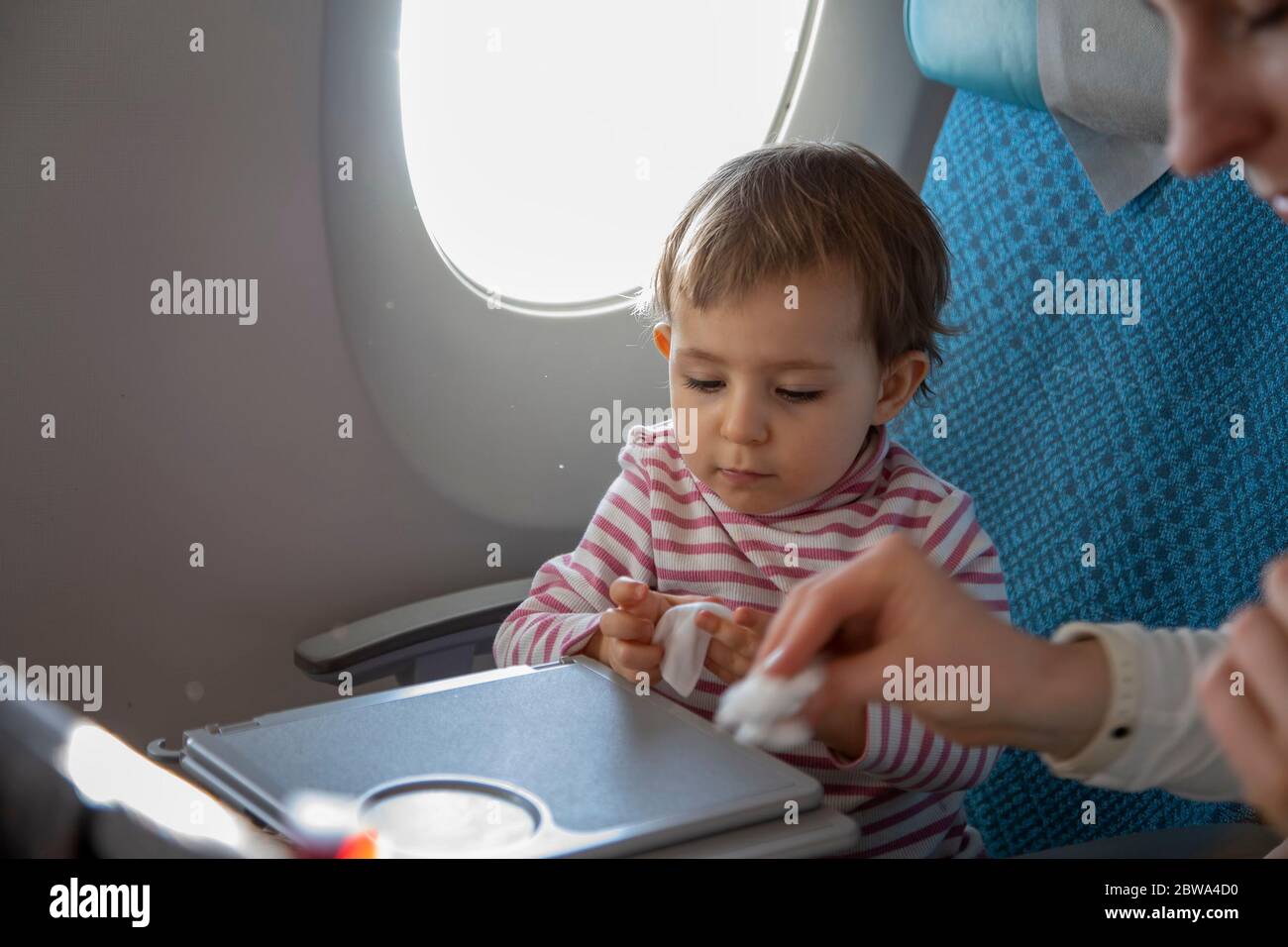 Ein kleines süßes Kleinkind Mädchen sitzt in einem Flugzeug in einem Stuhl am Bullauge und nimmt ein nasses antibakterielles Tuch von ihrer Mutter, um ihren Tisch abzuwischen Stockfoto