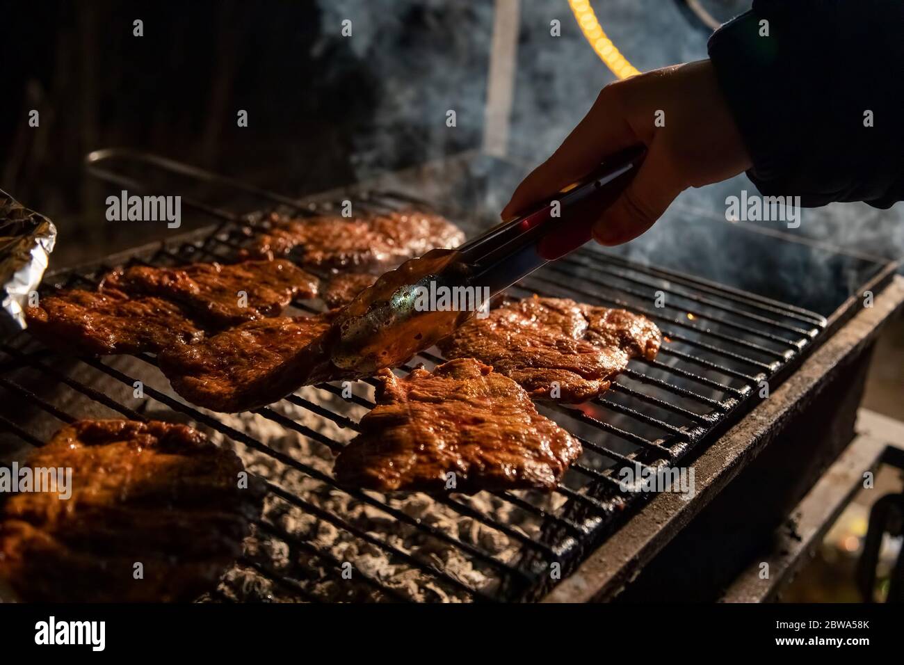 Abends ein Barbecue-Grill, auf dem leckere saftige Steaks über offenem Feuer gegrillt werden, eine Hand hält die Zange und dreht das Fleisch um. Nahaufnahme, weich Stockfoto