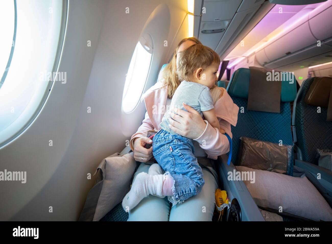 Konzept des Fluges mit einem Kind. Kleines niedliches Kleinkind, das auf den Knien springt, mit einer jungen schönen Mutter in einem Flugzeugsessel Stockfoto