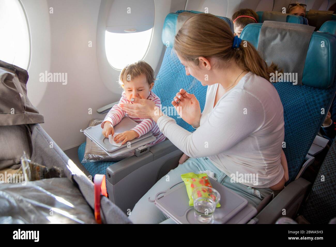 Kleines süßes Kleinkind Mädchen sitzt in einem Flugzeug in einem Stuhl am Bullauge, hält ein nasses Handtuch in den Händen und niest. Mama sitzt in der Nähe und bedeckt sie Stockfoto