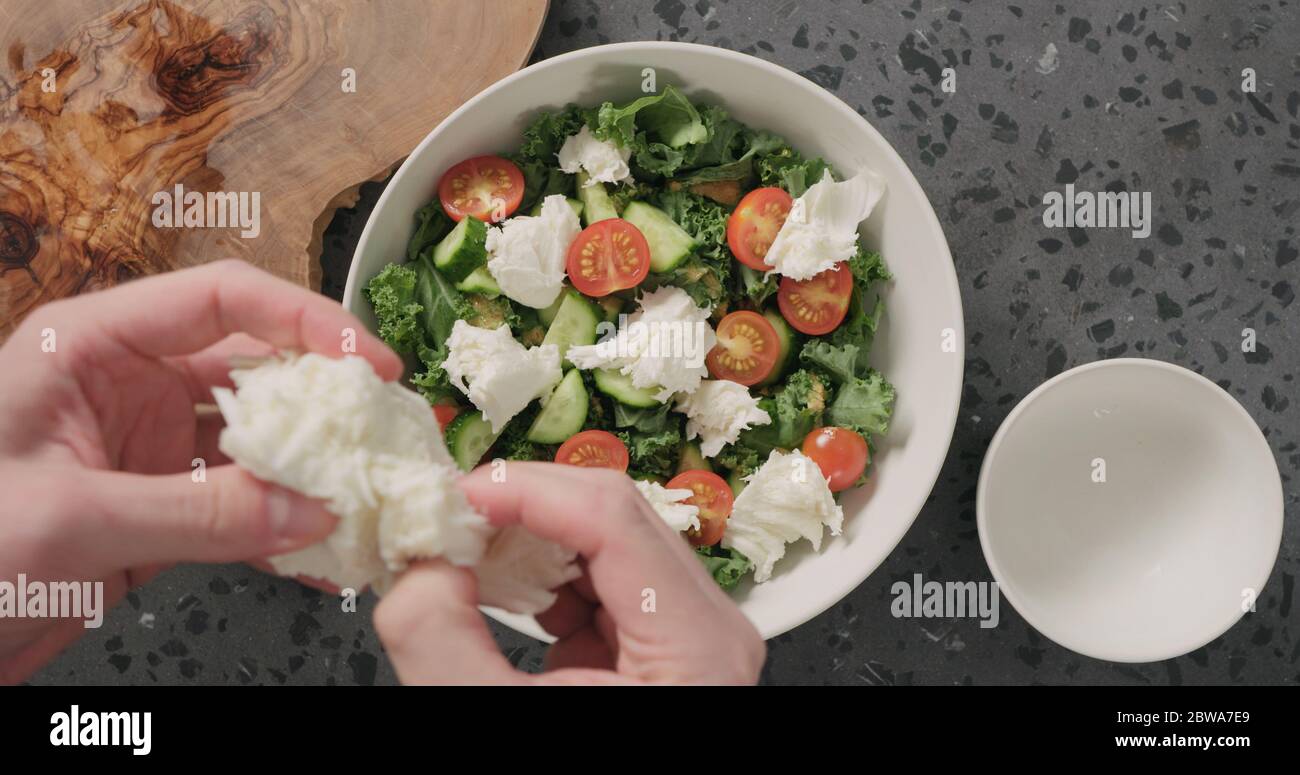 Draufsicht man Hands fügen Mozzarella über Grünkohlensauce und Tomaten In weißer Keramikschale auf Betongrund Stockfoto