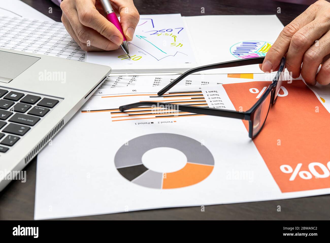 Detail der Person Hände Zeichnung Grafiken auf einem Papier und halten Brille mit der anderen Hand von einem Laptop und Papier mit Zahlen und Grafiken umgeben Stockfoto