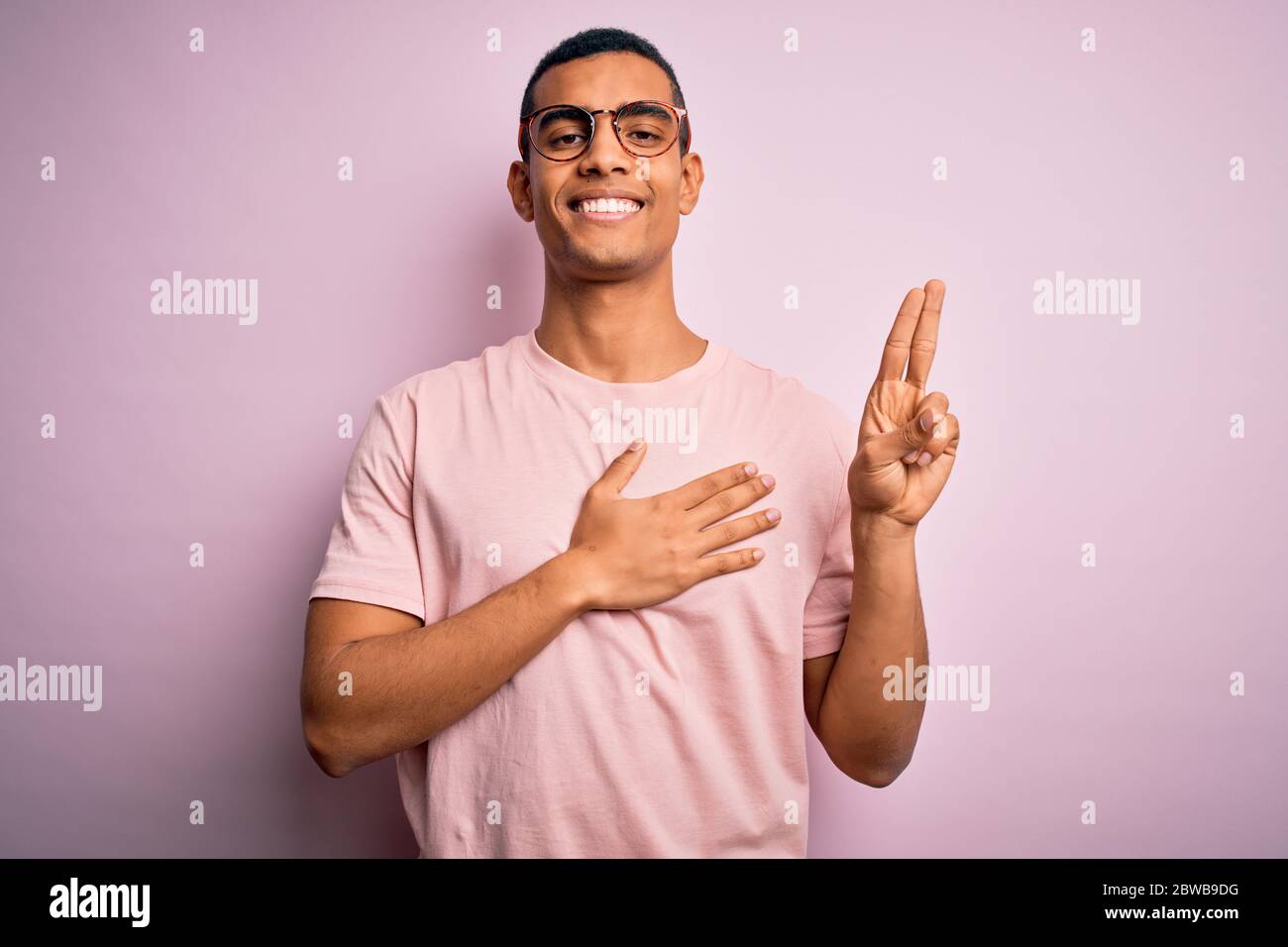 Schöner afroamerikanischer Mann, der lässiges T-Shirt und Brille auf rosa Hintergrund trägt und lächelnd mit der Hand auf der Brust und den Fingern nach oben flucht, was einen loy macht Stockfoto