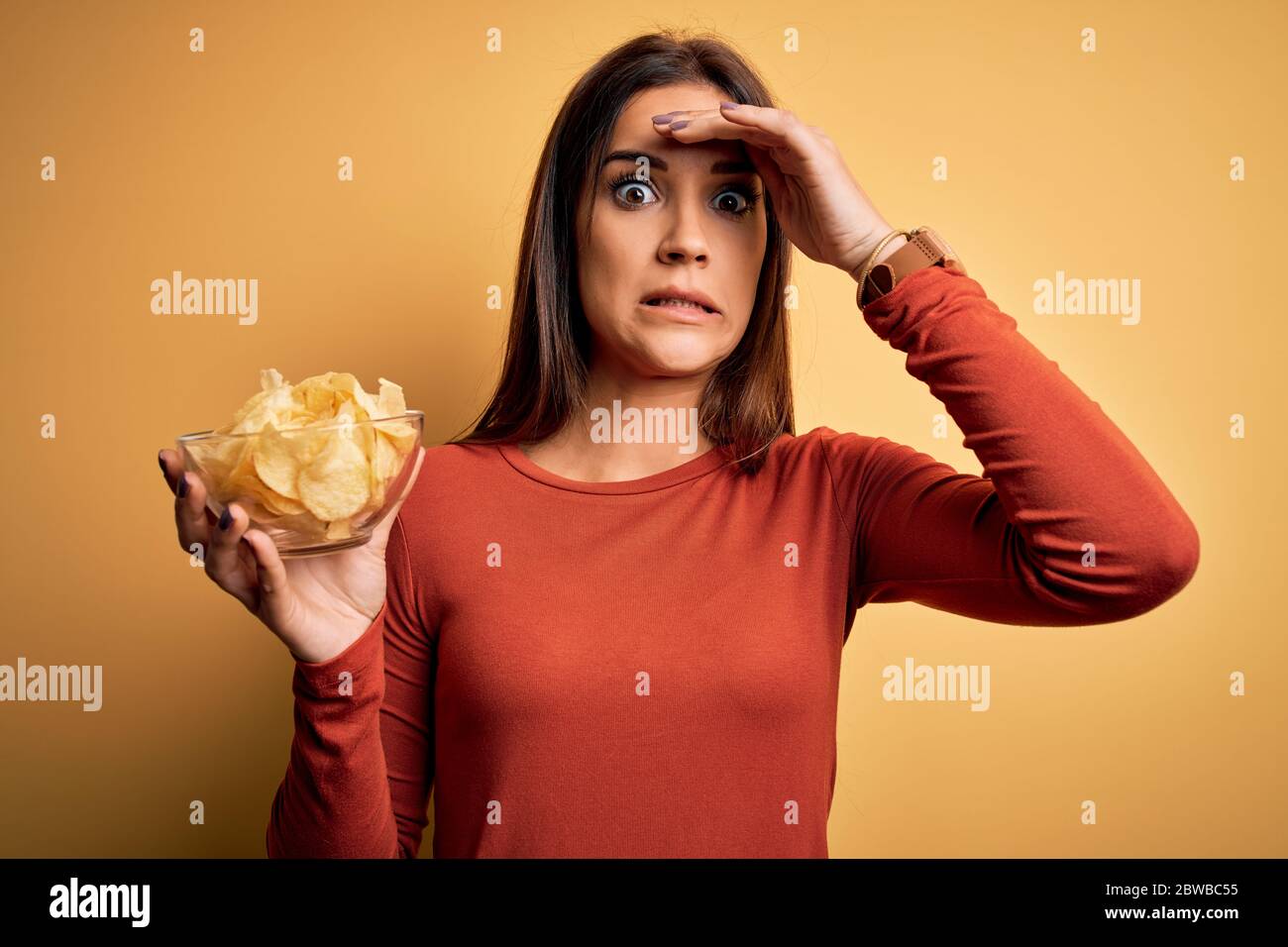 Junge schöne Brünette Frau hält Schüssel mit Chips Kartoffeln über gelbem Hintergrund mit Hand auf Kopf gestresst, schockiert mit Scham und Überraschung fac Stockfoto