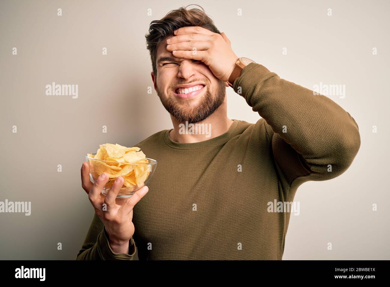 Junge blonde Mann mit Bart und blauen Augen hält Schüssel mit ungesunden Kartoffeln Chips gestresst mit Hand auf den Kopf, schockiert von Scham und Überraschung Gesicht, Stockfoto