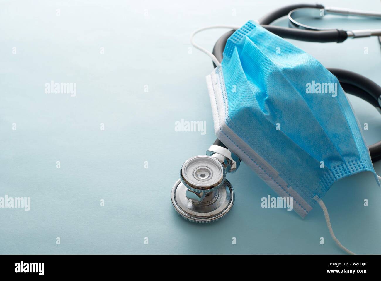 Chirurgische Gesichtsmaske drapiert über ein Stethoskop, das hoch angeschaut wird, über einem weißen Hintergrund mit Kopierraum konzeptuell der Covid-19 Pandemie, Gesundheitswesen Stockfoto