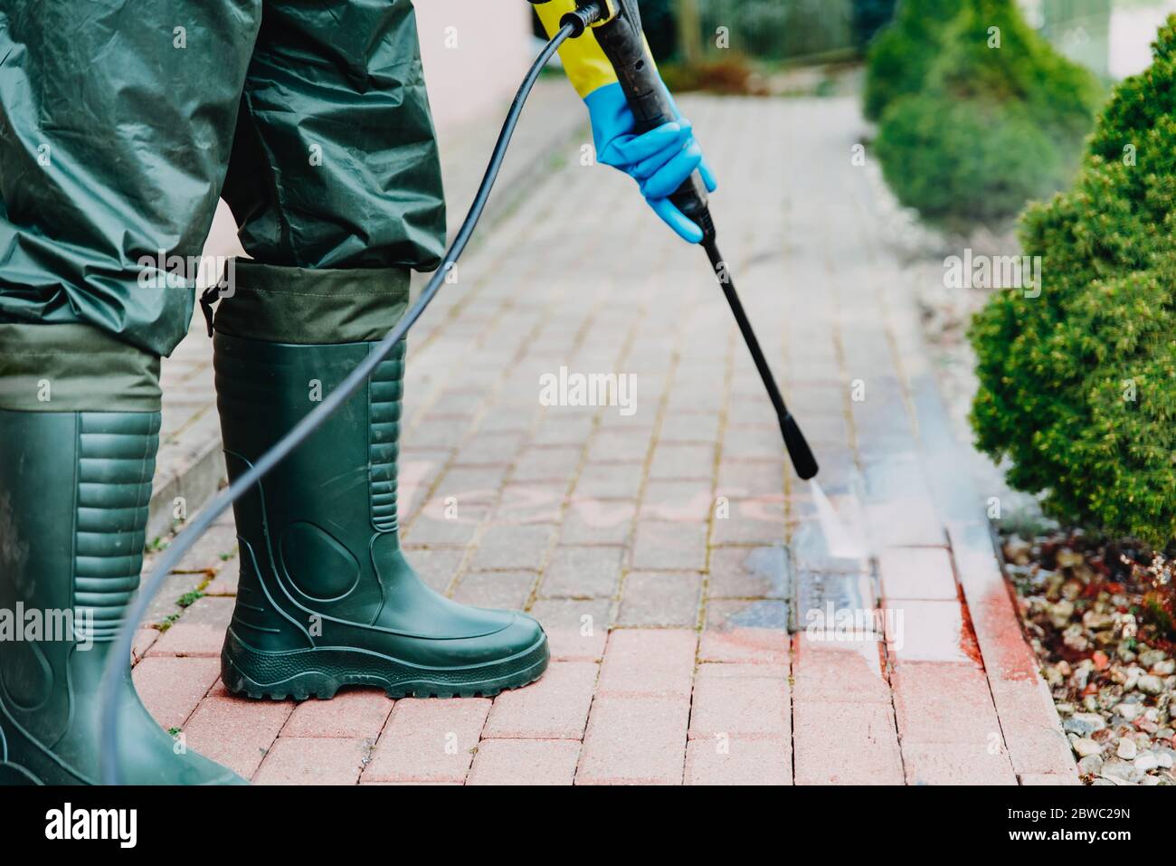 Mann putzt roten, conrete Bürgersteig Block mit Hochdruck-Wasserdeck. Reinigungskonzept für Pflaster. Mann mit Wathosen, wasserdichte Schutzhose Stockfoto