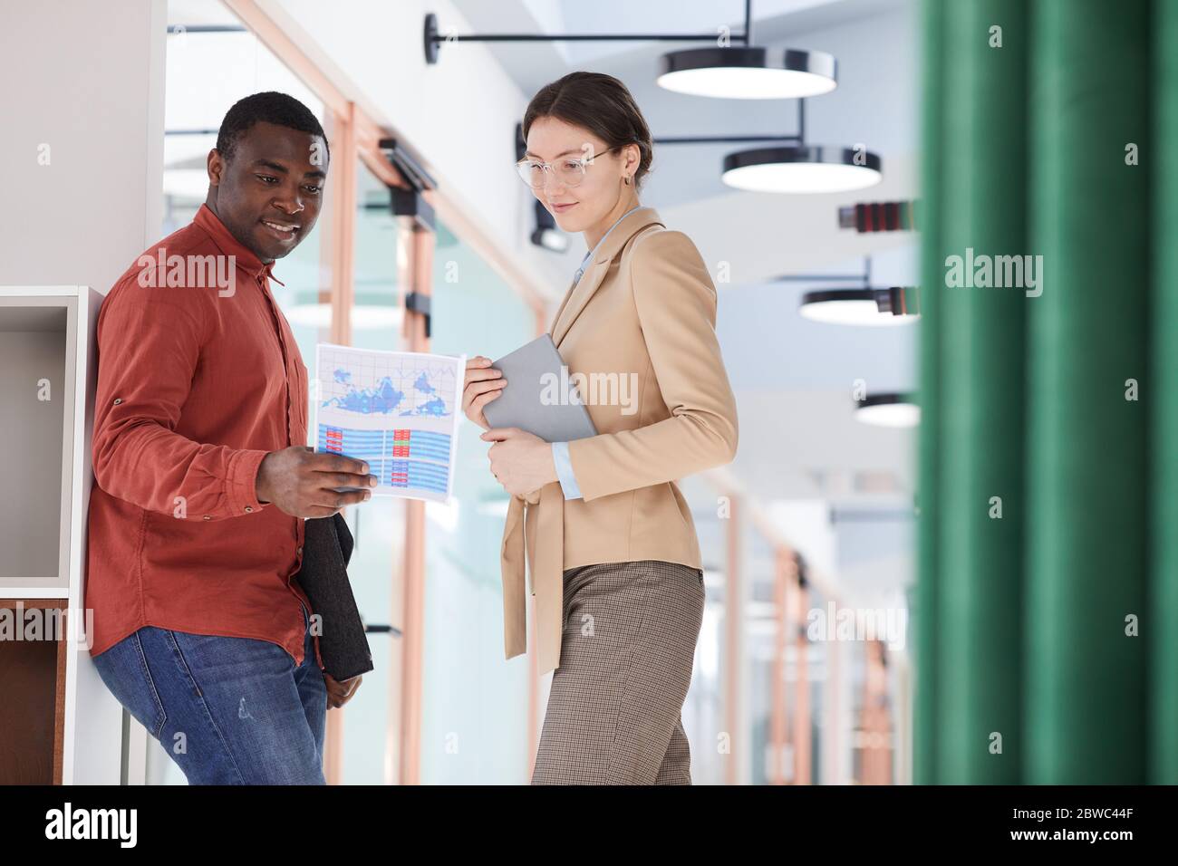 Porträt eines lächelnden afroamerikanischen Mannes, der Dokumente hält und mit einer eleganten jungen Geschäftsfrau im zeitgenössischen Bürogebäude spricht Stockfoto
