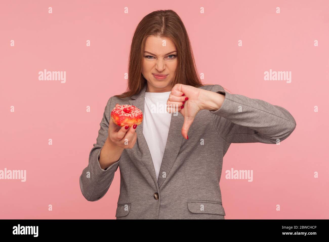 Porträt der unzufriedenen Frau Mitarbeiter in Anzug Jacke mit süßen Donut und zeigt Daumen nach unten, unzufrieden mit ungesunden Snack, Junk Food für Stockfoto