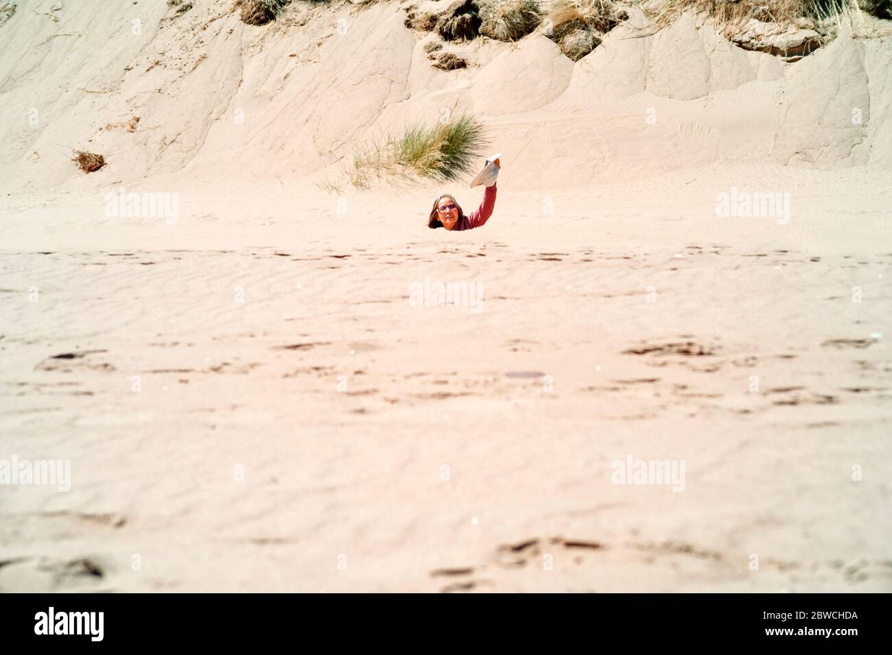 Fernbild der asiatischen Frau trägt Sonnenbrille winkenden Hut, bis zum Hals unter Sanddünen am Strand Konzept versenkt: Sinkende Gefühl, Treibsand. Stockfoto