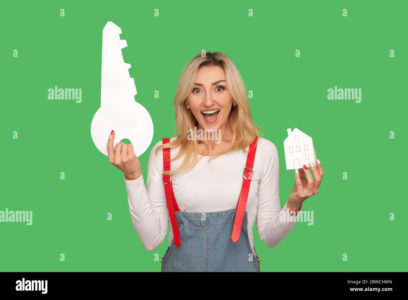 Porträt von aufgeregt blonde Frau in stilvollen Denim Overalls Anhebung großen Schlüssel und Papier Haus, schreiend vor Glück, Freude am Kauf von echten est Stockfoto
