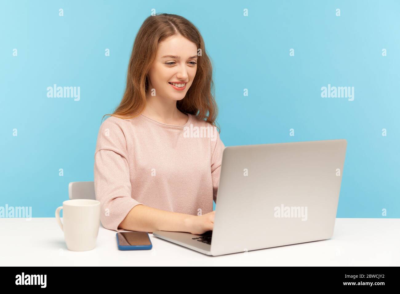 Fröhliche junge Geschäftsfrau in Freizeitkleidung sitzt am Arbeitsplatz, tippen auf Laptop und lächeln, online arbeiten, tun freiberufliche Arbeit im Home Office. Stockfoto