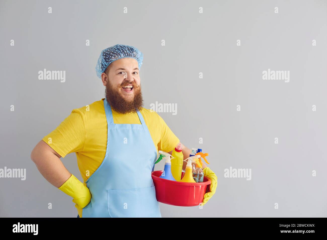 Lustige dicke Mann mit einem Bart in einer Schürze reinigt auf einem grauen Hintergrund. Stockfoto