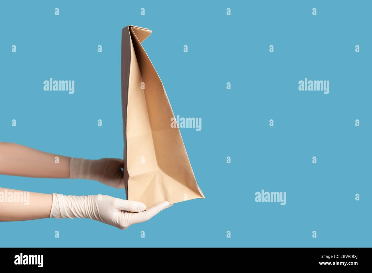 Profil Seitenansicht Nahaufnahme der menschlichen Hand in weißen chirurgischen Handschuhen halten und Handwerk Papierpaket mit Lebensmitteln. Sichere Lebensmittel-Delivery-Konzept. Indoor, Studio Stockfoto