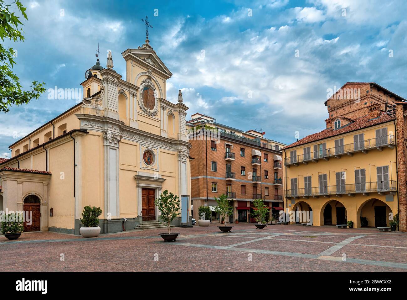 Katholische Kirche und historisches Haus auf dem kleinen Stadtplatz unter bewölktem Himmel in Alba, Piemont, Norditalien. Stockfoto