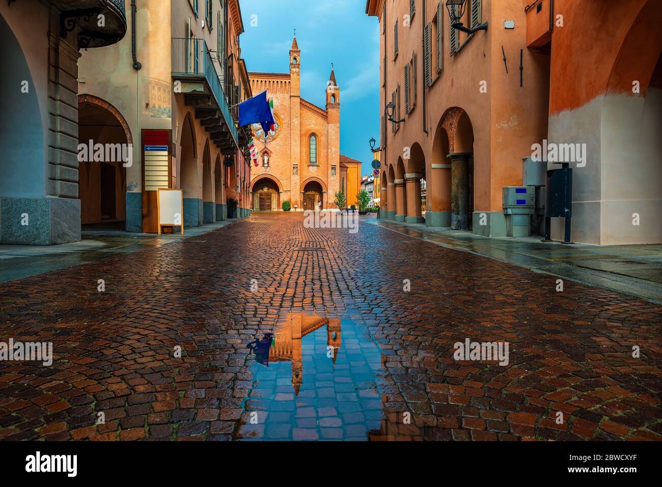 Kleine Pfütze auf schmalen Kopfsteinpflasterstraße zwischen alten Häusern und San Lorenzo Kathedrale im Hintergrund in der Stadt Alba, Piemont, Norditalien. Stockfoto